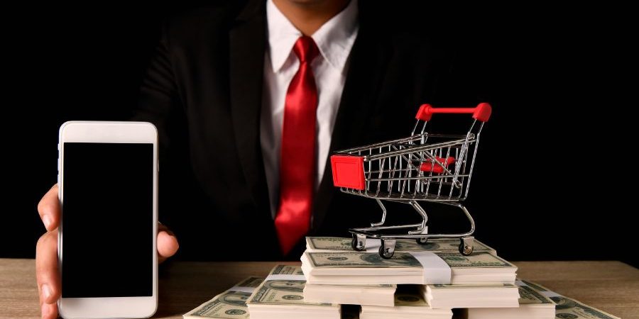 Midsection of businessman showing smart phone by paper currencies and shopping cart at table against black background